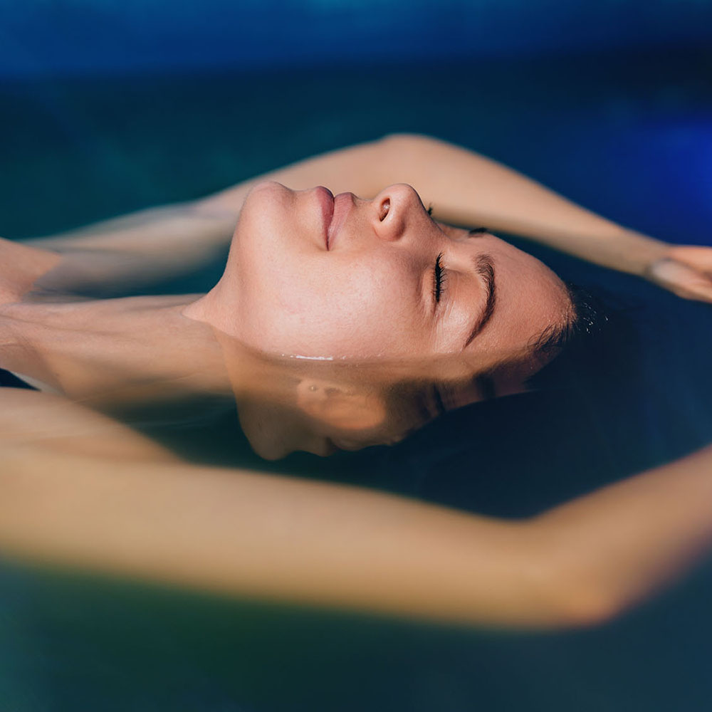 Woman floating in sensory deprivation tank with blue ambient lighting.